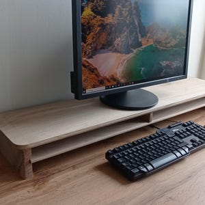 Op de afbeelding: Een lichtgekleurde houten monitorstandaard met een zwarte computermonitor. Een zwart toetsenbord staat op het bureau voor de standaard. De standaard heeft een onderste plank voor opslag, in cm.
