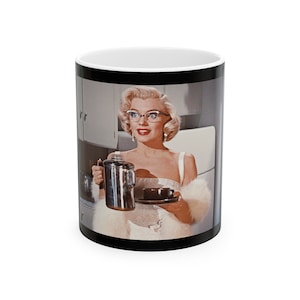 Puede incluir: Taza de café de cerámica blanca con una imagen vintage de una mujer rubia con gafas, sosteniendo una cafetera y una taza de café. La taza tiene una base negra y un interior blanco.
