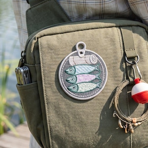 May include: An olive green canvas crossbody bag featuring a circular embroidered patch of a sardine tin with three fish. A metal carabiner holds a coiled fishing line and a red and white bobber. A multi-tool is tucked into the side pocket.