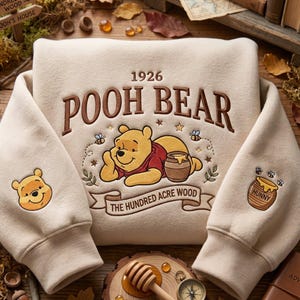 Op de afbeelding: Een beige sweatshirt met een geborduurde afbeelding van Winnie de Poeh met een honingpot, de tekst 1926 POOH BEAR en THE HUNDRED ACRE WOOD op een banner. De mouwen tonen een gezicht van Poeh aan de ene kant en een honingpot aan de andere kant.