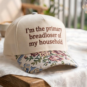 Op de afbeelding: Een beige baseballpet met de geborduurde tekst "I'm the primary breadloser of my household." De klep heeft een bloemenpatroon in blauw, groen en roze. De pet ligt op een houten tafel.