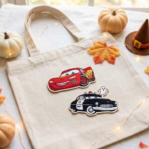 May include: A natural-colored canvas tote bag featuring two embroidered patches: a red race car with the number 95 and a black police car with a small white ghost figure on its roof. The bag has two fabric handles and a simple rectangular shape.
