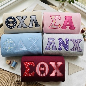 Peut inclure: Cinq sweat-shirts pliés de couleur crème, rose, bleu clair, gris et bordeaux, chacun orné de grandes lettres grecques appliquées dans divers motifs incluant des étoiles, du vichy, des pois et des imprimés floraux, sur une surface tressée.