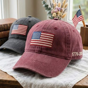 May include: Two baseball caps, one gray and one burgundy, each featuring an embroidered American flag patch. The burgundy cap has "1776-2006" printed on the side. Both caps are sitting on a wooden surface.