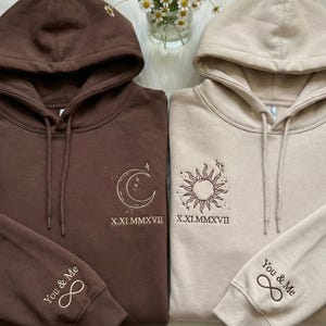 May include: Two matching hoodies, one brown and one beige, with embroidered celestial designs. The brown hoodie features a moon and stars, while the beige one has a sun. Both have the text "You & Me" and an infinity symbol on the sleeve.