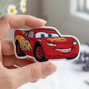 May include: An embroidered patch featuring a red race car with blue eyes, a white smile, and the number 95 in yellow on the side. The car has black wheels and a yellow lightning bolt graphic. The patch has a white border and detailed stitching.