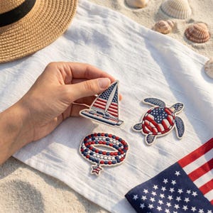 Op de afbeelding: Drie geborduurde patches met patriottische thema's: een zeilboot met een zeil in de stijl van de Amerikaanse vlag, een zeeschildpad met een schild in vlagpatroon en een kralenarmband met een kleine stervormige hanger in rood, wit en blauw.