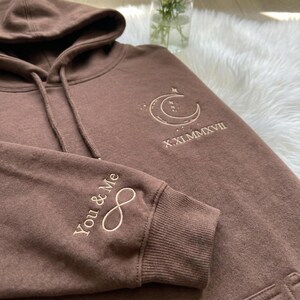 May include: Brown hoodie with a drawstring hood and a kangaroo pocket. The left sleeve features the embroidered text "You & Me" with an infinity symbol. The right chest has an embroidered crescent moon and stars design with the Roman numeral date X.XI.MMXVII.