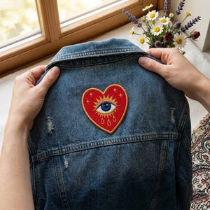May include: A blue denim jacket featuring an embroidered patch on the back. The patch is a red heart shape with a central blue eye, gold sunburst rays, and small gold teardrops and stars. The jacket has a classic collar and distressed texture.