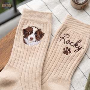 May include: A pair of beige ribbed socks. One sock features an embroidered portrait of a brown and white dog. The other sock features the embroidered name Rocky in brown script lettering above a small brown embroidered dog paw print.