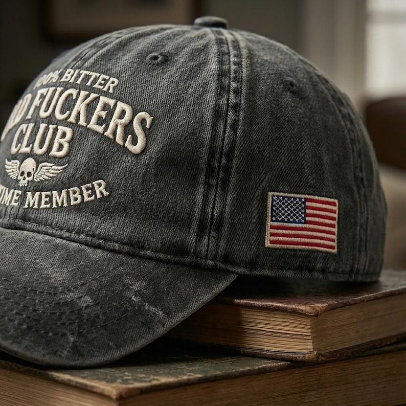 May include: A faded black baseball cap with white embroidered text reading "100% Bitter, Good Fuckers Club, Lifetime Member". It has a skull and wings emblem and a small American flag patch. The cap rests on a pile of aged books.