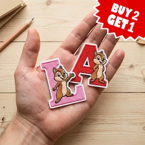 May include: Two embroidered patches shaped like the letters L and A, each featuring a brown chipmunk character. The L patch is light pink with a white border, and the A patch is red with a white border. The chipmunks have tan bellies and dark brown stripes.