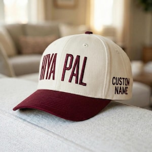 May include: A cream and maroon baseball cap with embroidered text. The front of the cap displays the words "NIYA PAL" in maroon, with "CUSTOM NAME" embroidered on the side. The cap is resting on a light-coloured surface.