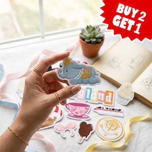 May include: An embroidered patch featuring a light blue elephant with a yellow and teal saddle. The patch is held up, surrounded by other assorted embroidered patches including a teacup, a monorail, a popsicle, and a circular emblem.