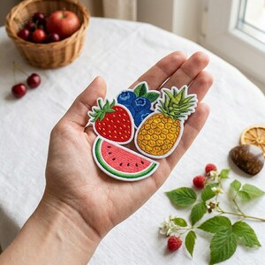 Op de afbeelding: Een set van vier geborduurde fruitpatches bestaande uit een rode aardbei met groene blaadjes, een tros blauwe bosbessen met groene blaadjes, een gele ananas met groene blaadjes en een roze watermeloenpartje met een groene schil.