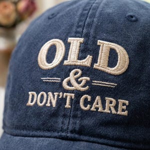 May include: A navy blue baseball cap with the embroidered text "OLD & DON'T CARE" in a light beige color. The cap is made of a soft fabric and has a curved brim.
