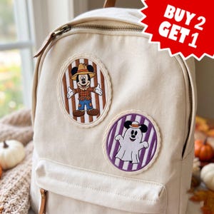 May include: A cream-colored canvas backpack featuring two circular patches. The top patch shows a scarecrow Mickey Mouse on a brown striped background. The bottom patch shows a ghost Mickey Mouse on a purple striped background. Text reads BUY 2 GET 1.
