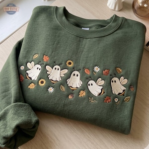 May include: An olive green crewneck sweatshirt featuring embroidered designs of five bees wearing white ghost costumes. The embroidery includes small sunflowers, white daisies, brown acorns, and scattered orange and brown autumn leaves.