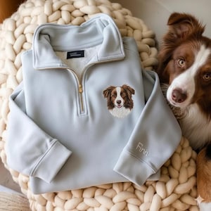 May include: A light blue pullover sweatshirt with a gold zipper and an embroidered dog portrait. The dog is brown and white. The name "Finn" is embroidered on the sleeve.