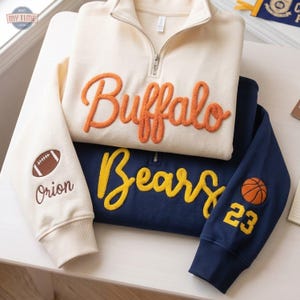 Può includere: Una felpa color crema con zip corta, scritta Buffalo in ciniglia arancione e un pallone da football ricamato con il nome Orion sulla manica, sovrapposta a una felpa blu navy con scritta Bears in ciniglia gialla.