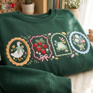 May include: A dark green crewneck sweatshirt featuring four embroidered rectangular and oval frames. The designs include a white goose with a green bow, a cluster of red strawberries with a pink bow, a green frog on a lily pad, and a floral bouquet.