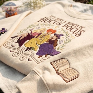May include: A cream-colored crewneck sweatshirt featuring an embroidered design of three figures in dresses and hats, with the text "IT'S JUST A BUNCH OF HOCUS POCUS" above them. An embroidered open book is on the sleeve, and a cat is on the shoulder.