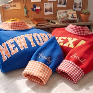 Puede incluir: Dos sudaderas de manga larga, una azul y otra roja, con cuellos altos de cuadros vichy en naranja y rojo. La sudadera azul muestra el texto NEW YORK, mientras que la roja indica TEXAS. Las mangas están bordadas con los nombres Emily y Sarah.