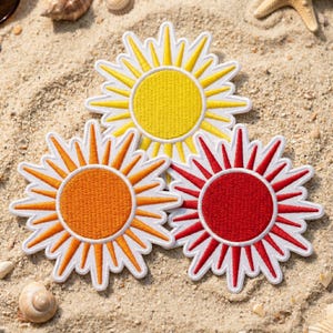 Op de afbeelding: Drie cirkelvormige geborduurde patches in de vorm van zonnen met stralende punten. Eén patch is felgeel, één is levendig oranje en één is dieprood. Elke zon heeft een massief cirkelvormig midden en een witte rand rondom de hele vorm.