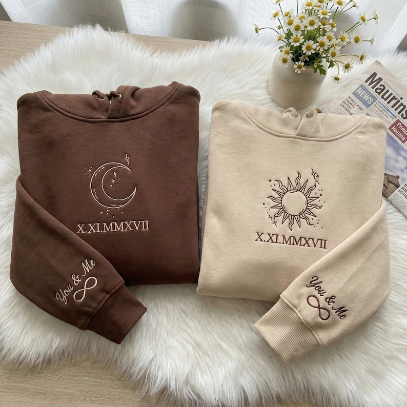 May include: Two hooded sweatshirts, one brown and one beige, displayed on a fluffy white surface. The brown sweatshirt features a crescent moon design, while the beige one has a sun graphic. Both include the text "X.XI.MMXVII" and "You & Me &infin;".