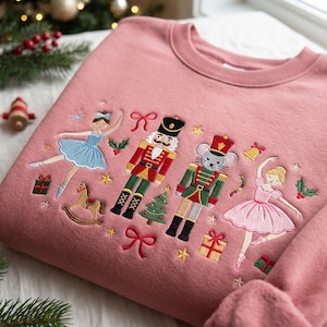 May include: A dusty rose crewneck sweatshirt featuring colorful embroidered holiday motifs including two ballerinas in blue and pink dresses, a nutcracker, a mouse soldier, a rocking horse, a Christmas tree, gift boxes, bows, and stars.