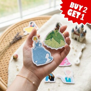 Op de afbeelding: Twee geborduurde stoffen patches met geanimeerde figuren in jurken, één in lichtblauw en één in groentinten. Een rode ster-afbeelding in de rechterbovenhoek bevat de witte tekst BUY 2 GET 1. Andere karakter-patches zijn op de achtergrond zichtbaar.