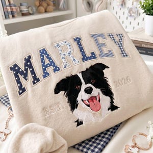May include: A beige sweatshirt featuring the name MARLEY in large, patterned fabric letters. Below the name is an embroidered portrait of a black and white dog with its tongue out, flanked by the text EST. and 2026 in beige thread.