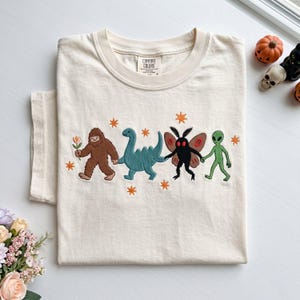 May include: A cream-colored t-shirt featuring an embroidered graphic of a brown Bigfoot holding a flower, a blue dinosaur, a black moth creature with red eyes, and a green alien holding hands, accented with small orange star-shaped embroidery.