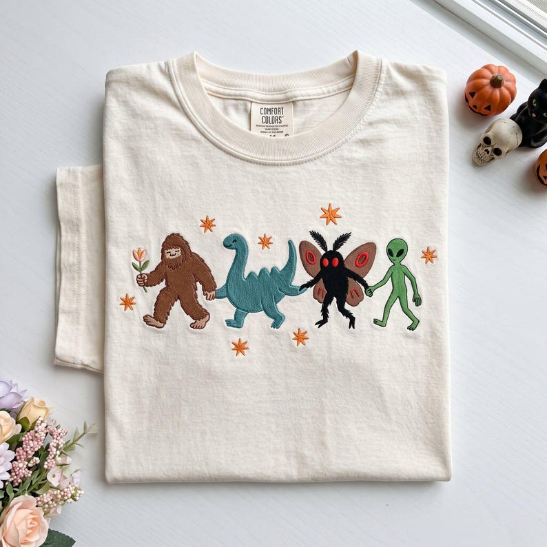 May include: A cream-colored t-shirt featuring an embroidered graphic of a brown Bigfoot holding a flower, a blue dinosaur, a black moth creature with red eyes, and a green alien holding hands, accented with small orange star-shaped embroidery.
