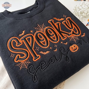 Może przedstawiać: Czarna bluza z okrągłym dekoltem z pomarańczowym haftowanym napisem "SPOOKY SEASON". Wzór zawiera pomarańczowe haftowane pajęczyny, małe nietoperze i gwiazdki otaczające napis, wraz z małą ikoną dyni umieszczoną przy tekście.