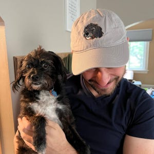 May include: A person wearing a grey baseball cap with an embroidered black and white dog on the front. The person is holding a black and white dog in their arms.