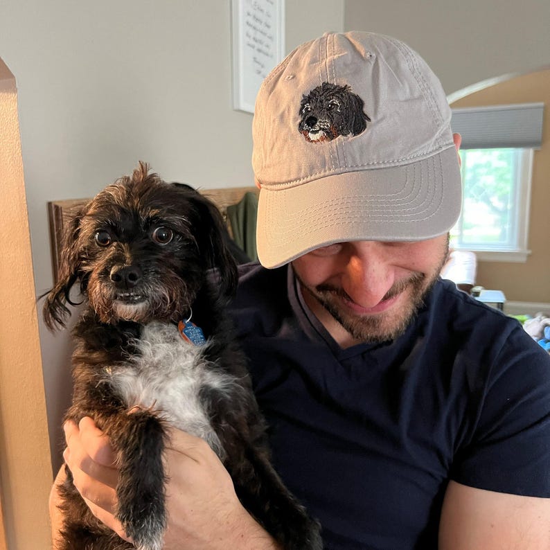 May include: A person wearing a grey baseball cap with an embroidered black and white dog on the front. The person is holding a black and white dog in their arms.