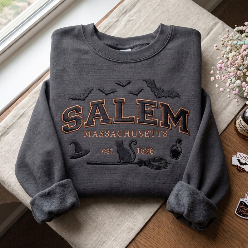 Op de afbeelding: Een antracietgrijze sweater met ronde hals en geborduurde tekst SALEM MASSACHUSETTS est 1626. Het ontwerp bevat zwarte silhouetten van vleermuizen, een heksenhoed, een kat, een bezemsteel en een toverdrankflesje in oranje en zwart garen.