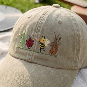 May include: A beige baseball cap featuring an embroidered design of four insects playing musical instruments. The embroidery depicts a green grasshopper with a trumpet, a red ladybug, a yellow and black bee with a trumpet, and a brown ant playing a cello.