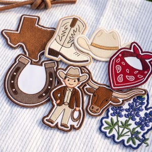 May include: A collection of embroidered patches featuring a brown Texas state shape, a cream cowboy boot, a cream cowboy hat, a brown horseshoe, a person in western attire, a brown longhorn steer head, a red paisley bandana, and blue flowers.