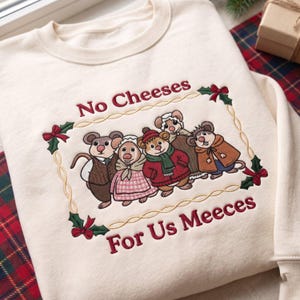Può includere: Una felpa color crema con un ricamo raffigurante cinque topi vestiti con abiti invernali. I topi sono incorniciati da un bordo a corda dorata con dettagli di agrifoglio. Il testo "No Cheeses For Us Meeces" è ricamato in rosso sopra e sotto.