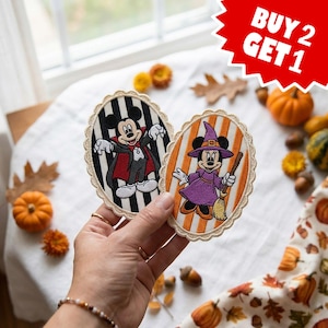 May include: Two oval-shaped embroidered patches featuring Disney characters. One patch shows Mickey Mouse as a vampire on a black and white striped background. The other shows Minnie Mouse as a witch on an orange and white striped background.