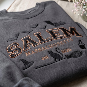 Op de afbeelding: Een antracietgrijze sweater met ronde hals en zwarte en koperkleurige borduursels. De tekst luidt SALEM MASSACHUSETTS est 1626. Het ontwerp bevat geborduurde vleermuizen, een heksenhoed, een zwarte kat, een bezemsteel en een klein flesje.