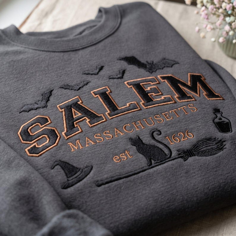 Op de afbeelding: Een antracietgrijze sweater met ronde hals en zwarte en koperkleurige borduursels. De tekst luidt SALEM MASSACHUSETTS est 1626. Het ontwerp bevat geborduurde vleermuizen, een heksenhoed, een zwarte kat, een bezemsteel en een klein flesje.