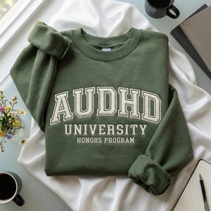 May include: An olive green sweatshirt featuring the text "AUDHD UNIVERSITY HONORS PROGRAM" in white, collegiate lettering. The sweatshirt is folded on a white cloth, with a coffee mug, flowers, and a notebook in the background.