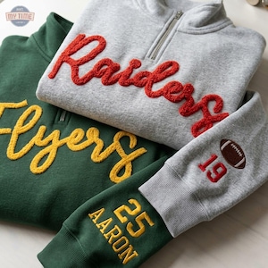 Può includere: Una felpa grigia con zip a un quarto, ricamo rosso Raiders e una manica con il numero 19 e un pallone da football, accanto a una felpa verde con ricamo giallo Flyers e una manica con il nome Aaron e il numero 25.