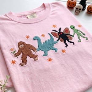 May include: A light pink crewneck t-shirt featuring an embroidered design of a brown Bigfoot holding a flower, a teal dinosaur, a black moth creature with red eyes, and a green alien holding hands, accented with small orange embroidered stars.