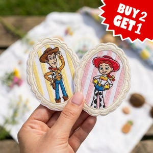 May include: Two oval embroidered patches featuring cartoon characters. One patch shows a cowboy with a brown hat and yellow striped background. The other shows a cowgirl with a red hat and pink striped background. Both have scalloped edges.
