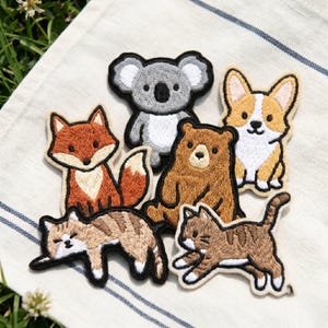 May include: A collection of six embroidered animal patches featuring a gray koala, an orange fox, a brown bear, a tan and white corgi, a light brown tabby cat lying down, and a brown tabby cat in a playful pouncing position with white accents.
