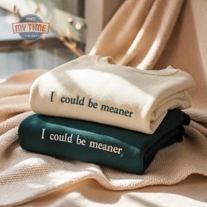 May include: Two folded sweatshirts, one cream-colored and one dark green, are stacked on top of each other. The cream sweatshirt has the embroidered text "I could be meaner" in a dark green font. The dark green sweatshirt also has the same text embroidered in white.